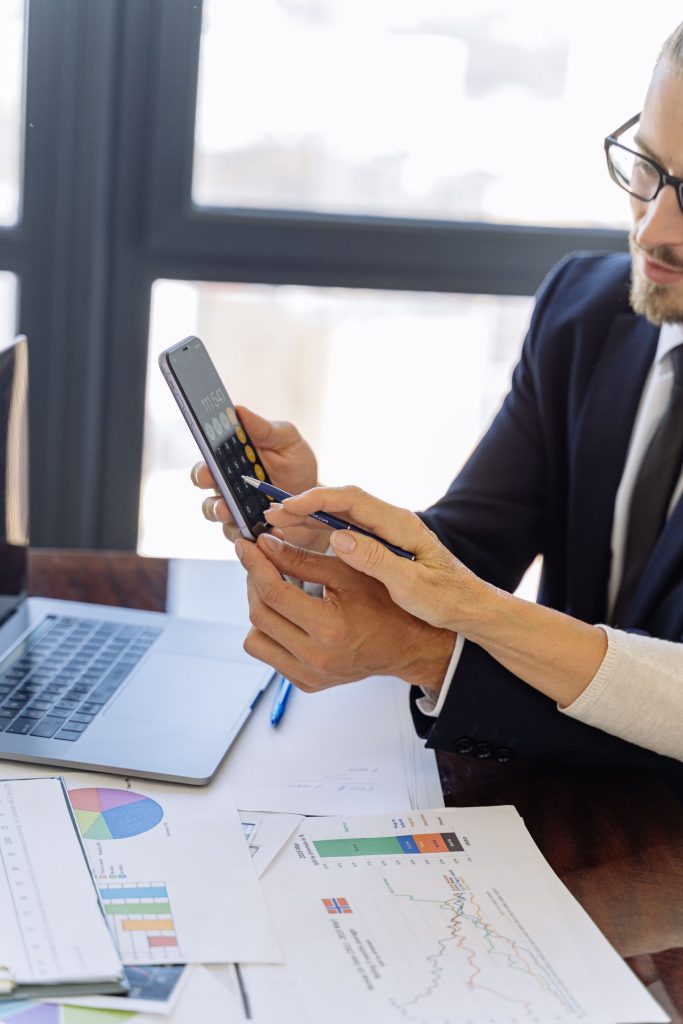 Business professionals reviewing financial reports on a smartphone at a desk with papers and a laptop in an office setting.